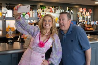 Monica (Sarah Hadland) stands in front of the bar at the rugby club, holding up her phone to film herself, with Terry (Johnny Vegas) standing behind her and joining in