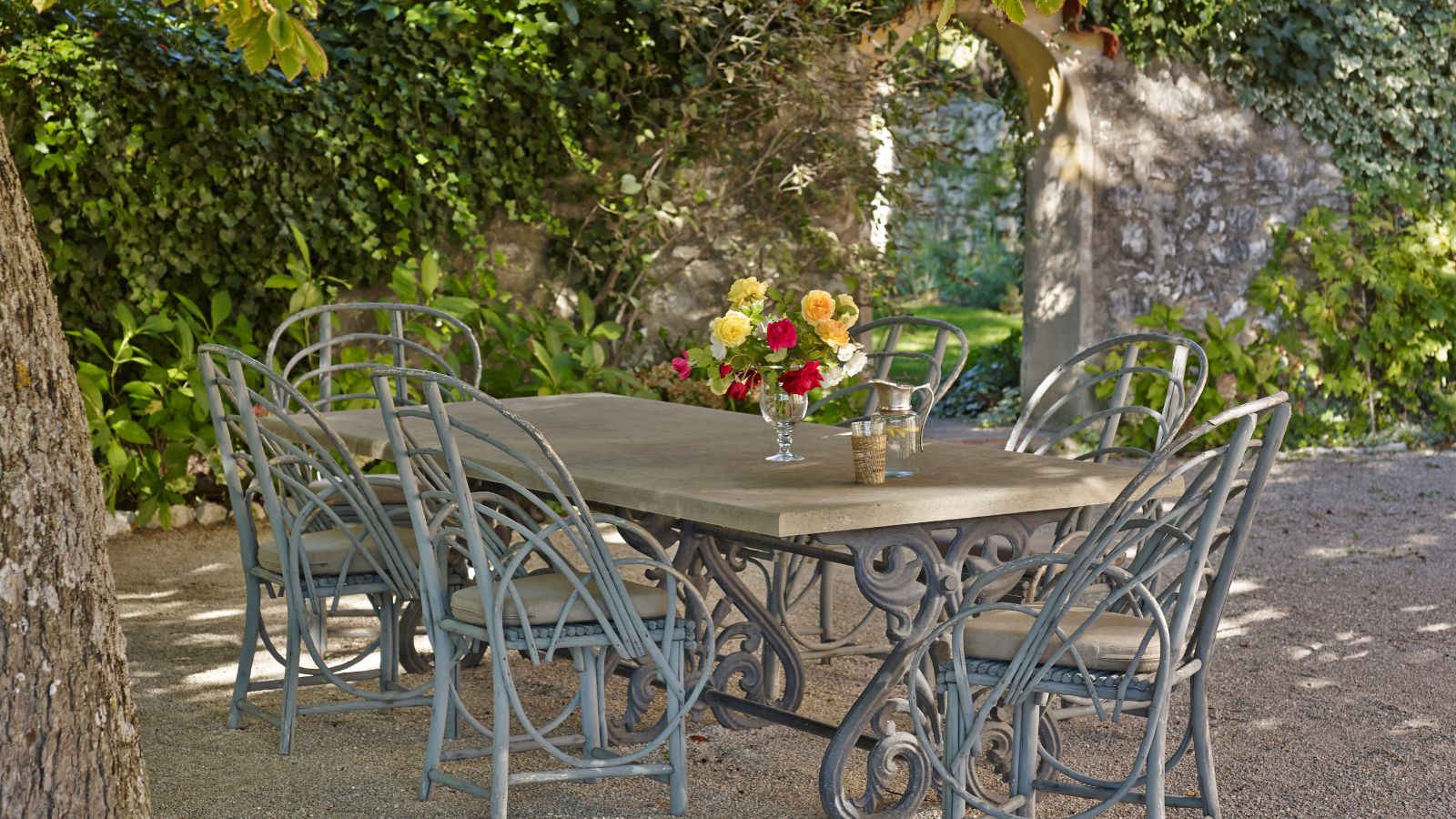 French garden with a brick arched way covered in ivy, and an antique wrought iron long dining table with vintage cane chairs underneath.