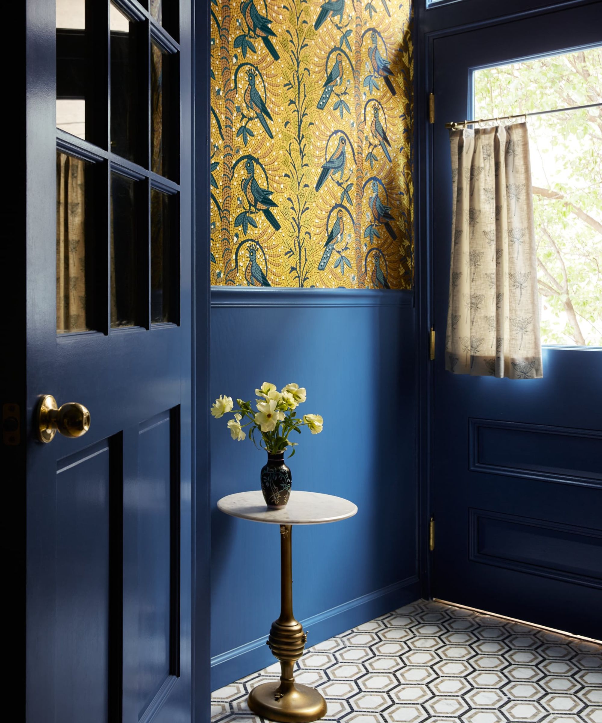 An entryway with dark blue panelling and doors, black and white geometric-tiled flooring, a small side table with a vase of yellow flowers, and yellow patterned wallpaper on the upper wall.