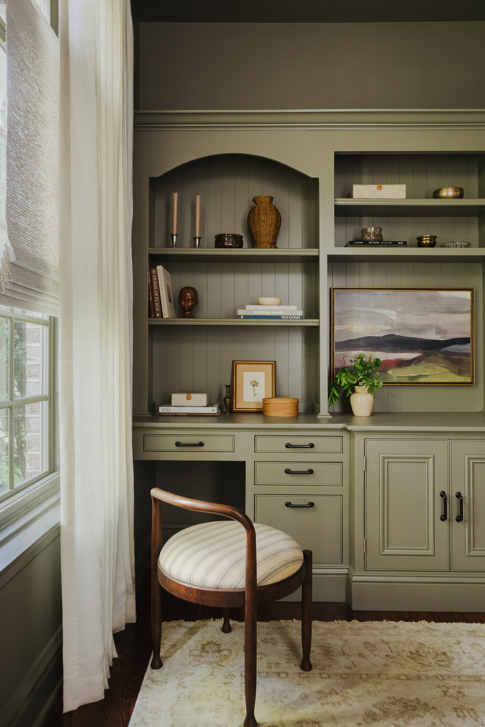 A sage green built-in bookshelf with a wooden and upholstered office chair. There are decorative objects on the bookshelf and a window with linen curtains.