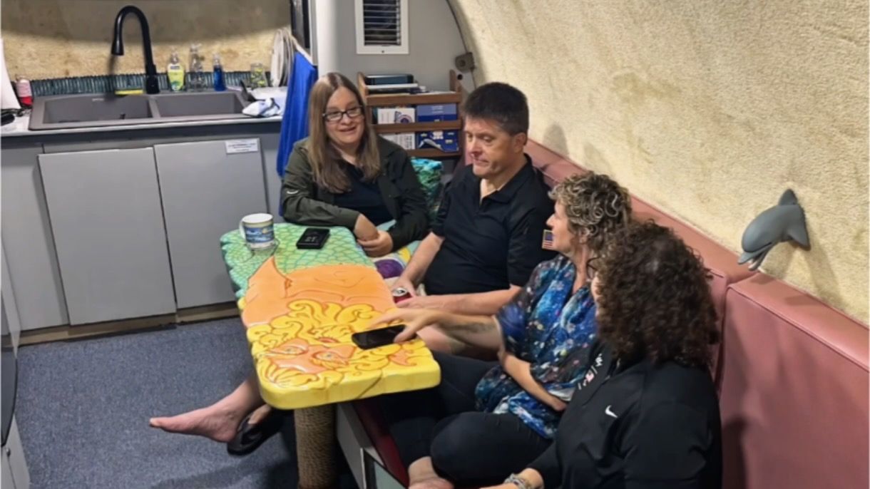 three women and one man sit on a red bench in a small kitchen with blue carpet