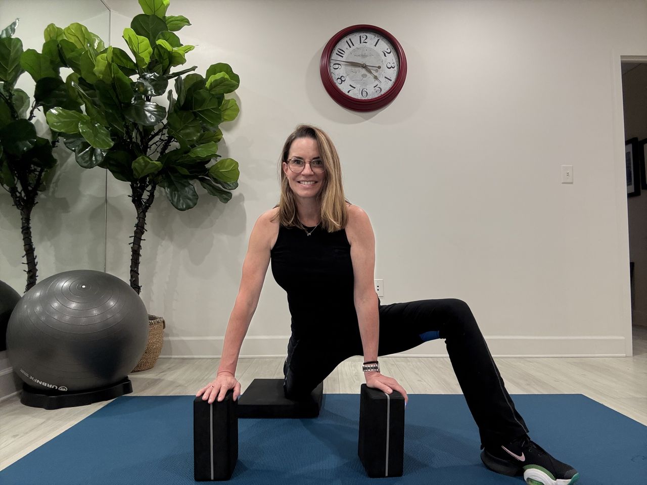 An employee at Femina physical therapy performs a kneeling lunge. Her front left leg is positioned out to the side as she leans forward, bending her left knee, to deepen the stretch.