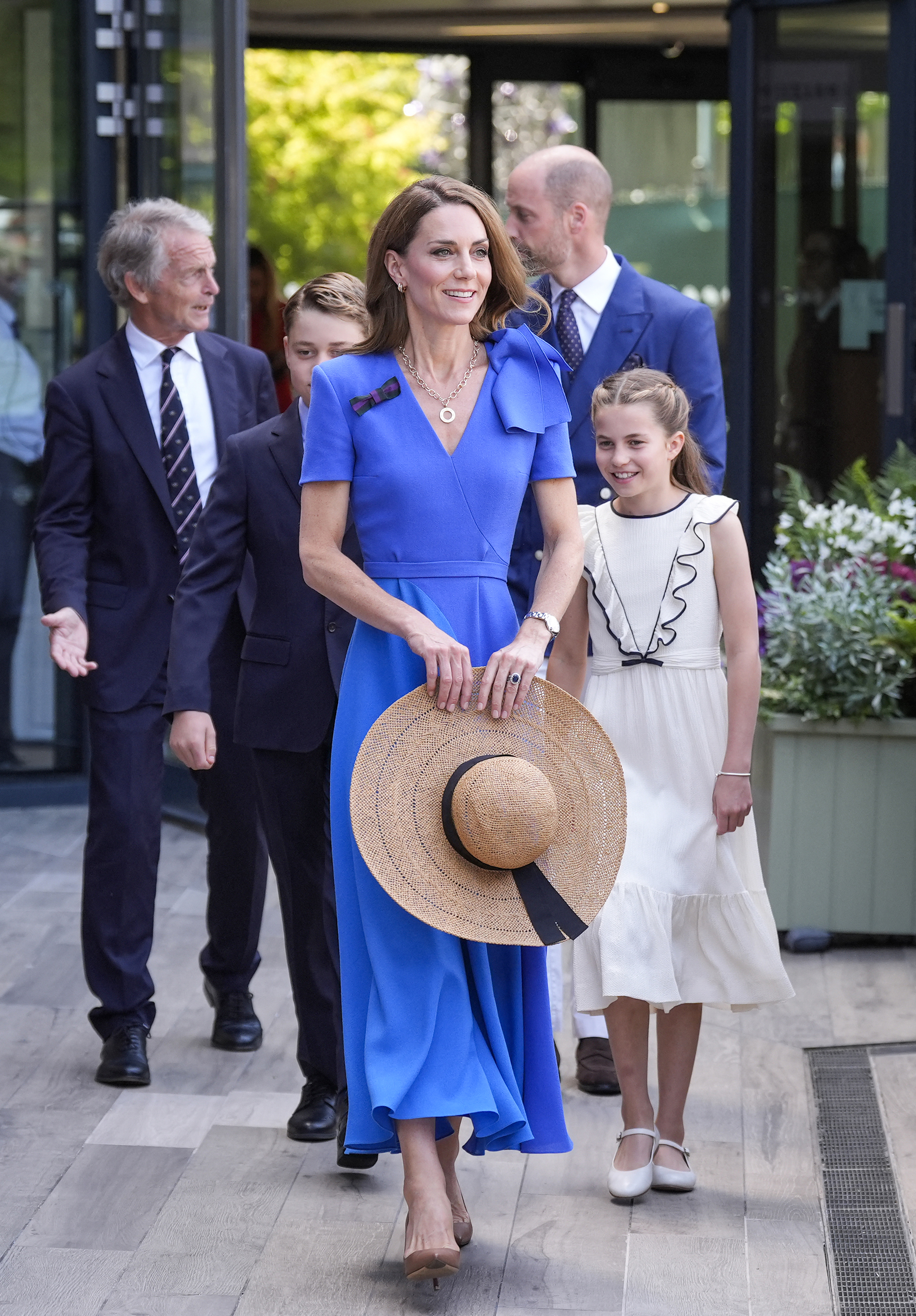 Princess Charlotte wearing a white dress walking behind Princess Kate in a blue dress and sun hat at Wimbledon
