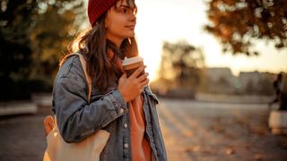 Woman wrapped up in a scarf and hat drinking a coffee outside in the morning sun