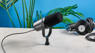 the shure mv7i, a black microphone with a black muff, touch LED pad, XLR connection and TRS input and USB-C input at the rear, photograhped against a blue tom's guide background with a pair of over-ear wired headphones 