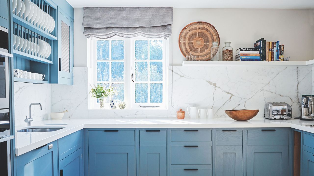 White marble kitchen with blue cabinets and a window directly in front with shelves to the right