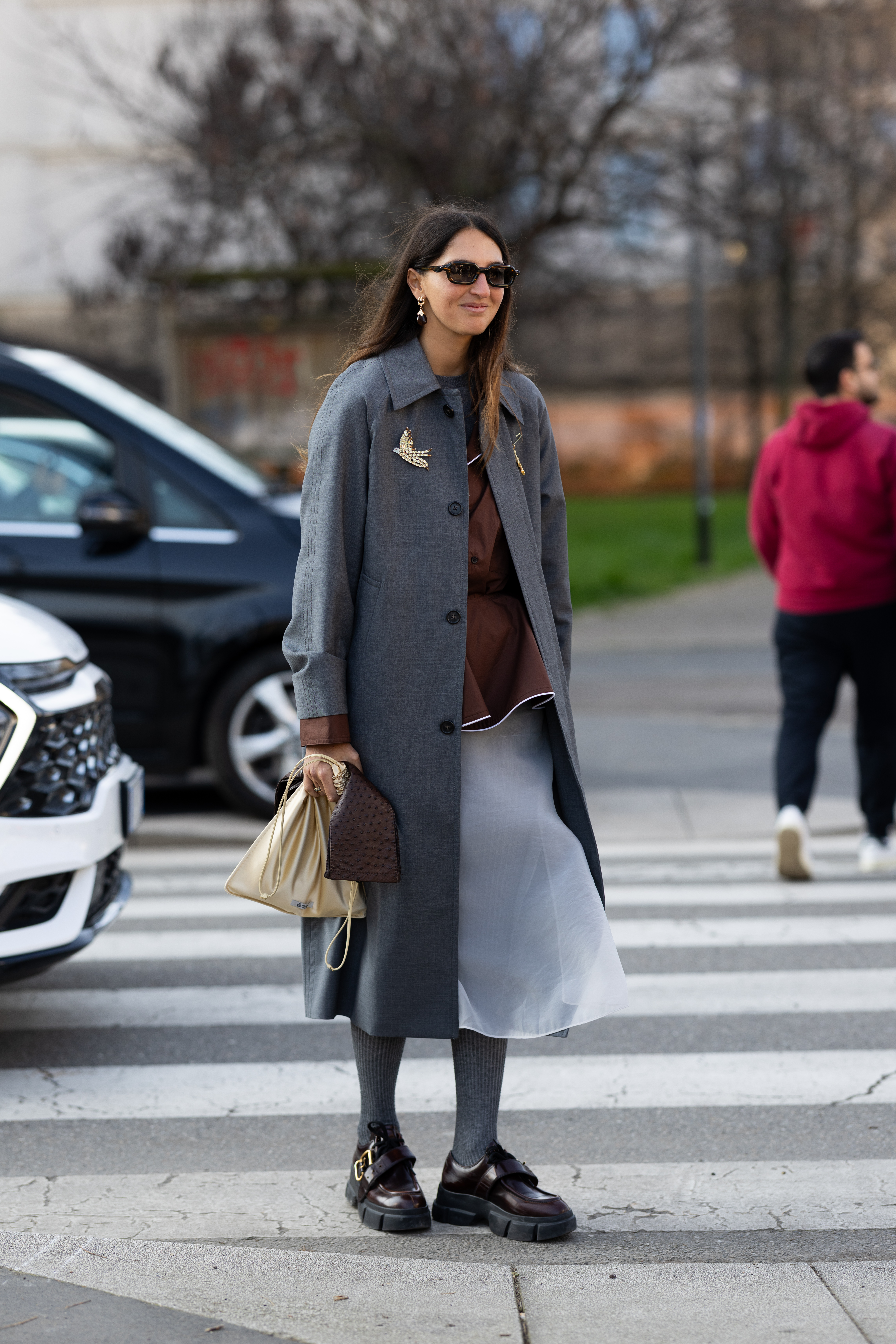 MILAN, ITALY - FEBRUARY 26: Marta Oldrini wears gold pins, black sunglasses, a grey coat, a brown shirt, a grey midi skirt, grey tights, brown leather loafers, a beige silk bag and a brown leather mini bag outside Prada show during the Milan Fashion Week - Womenswear Fall/Winter 2026/2027 on February 26, 2026 in Milan, Italy. (Photo by Valentina Frugiuele/Getty Images)
