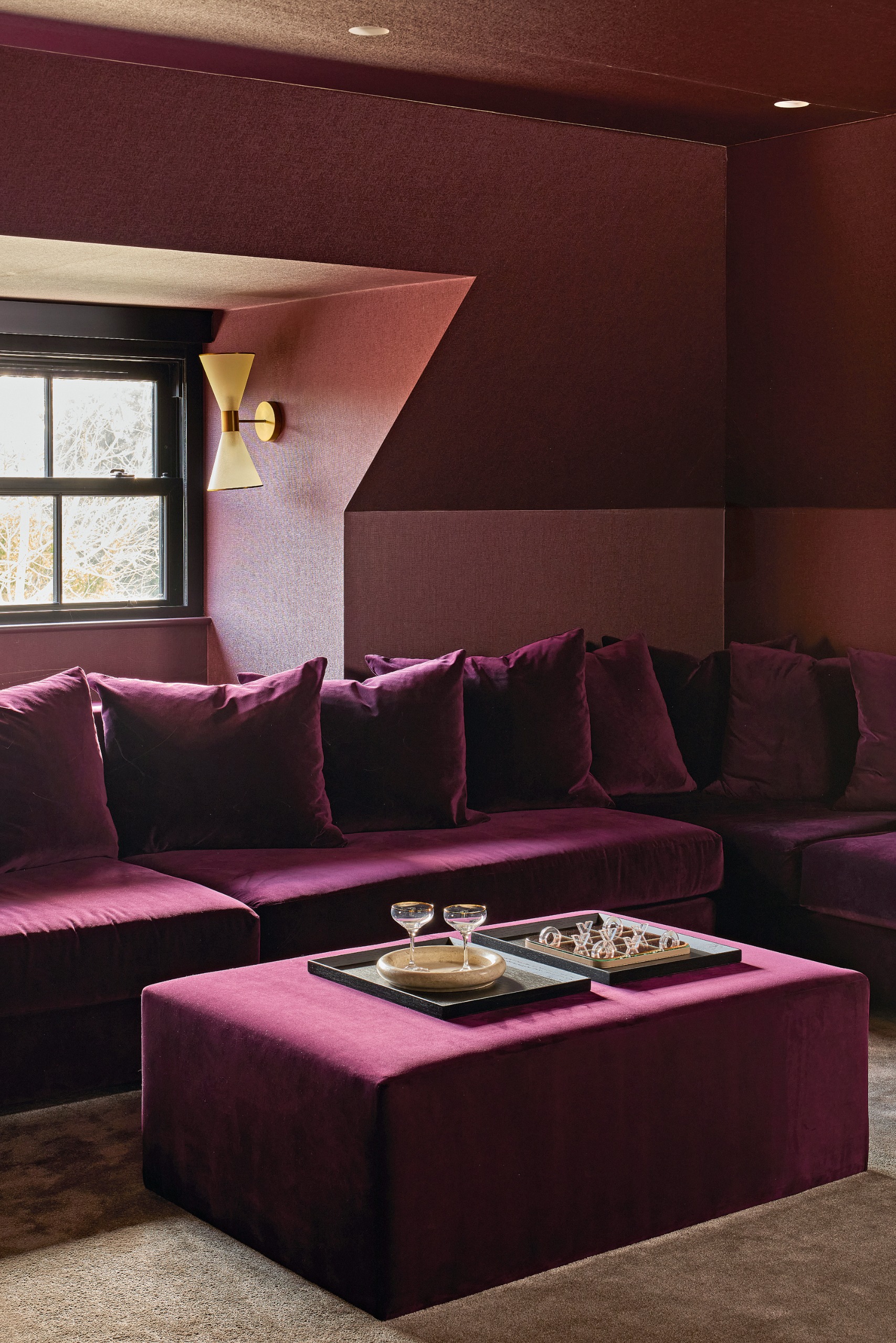 Cinema room with deep plum wallpaper, plum velvet corner sofa and ottoman, and brass wall light
