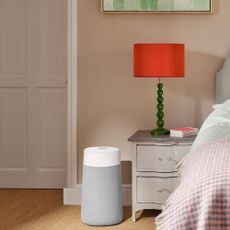 Blueair 3250i air purifier on the floor in a bedroom, next to a bedside table with a lamp on it