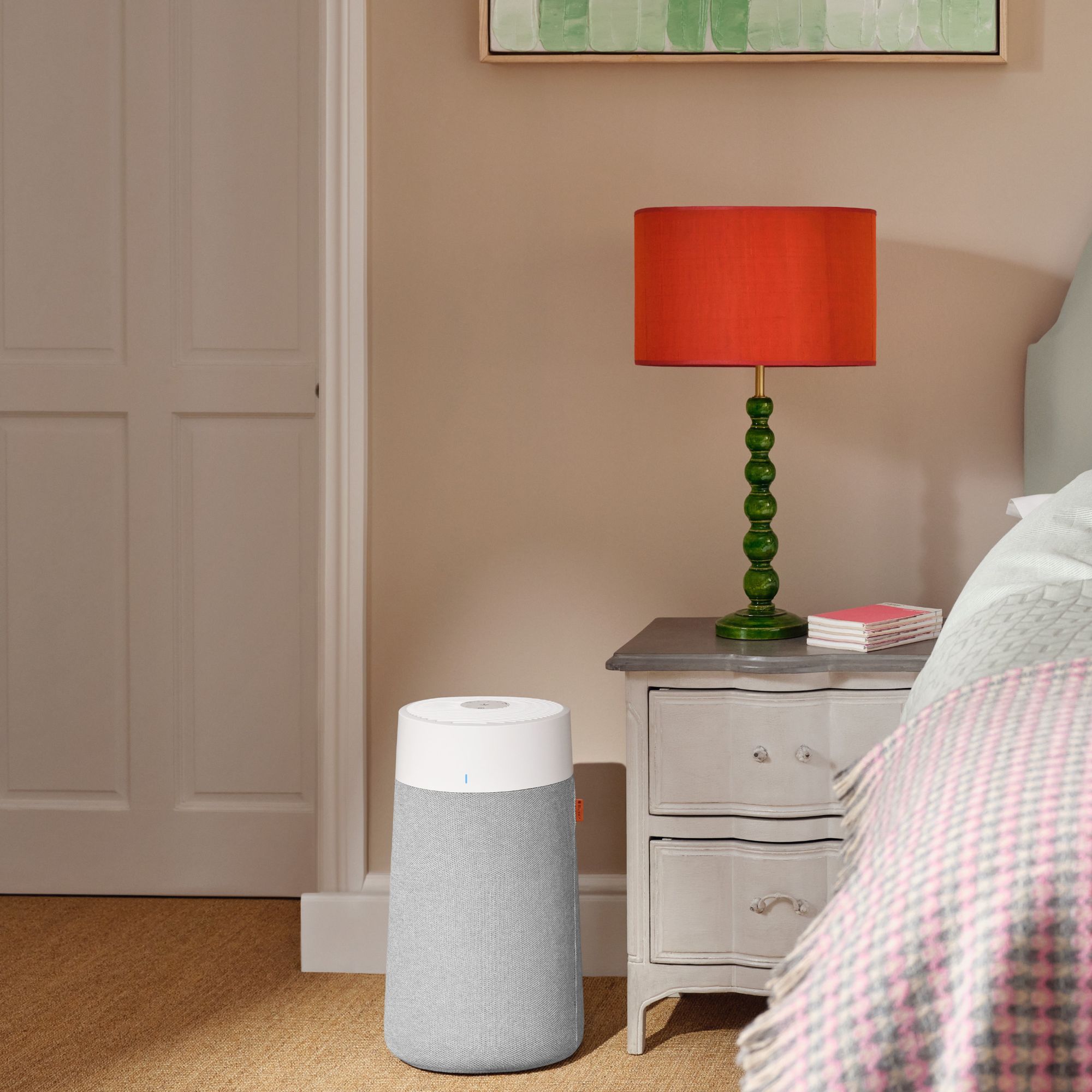 Blueair 3250i air purifier on the floor in a bedroom, next to a bedside table with a lamp on it