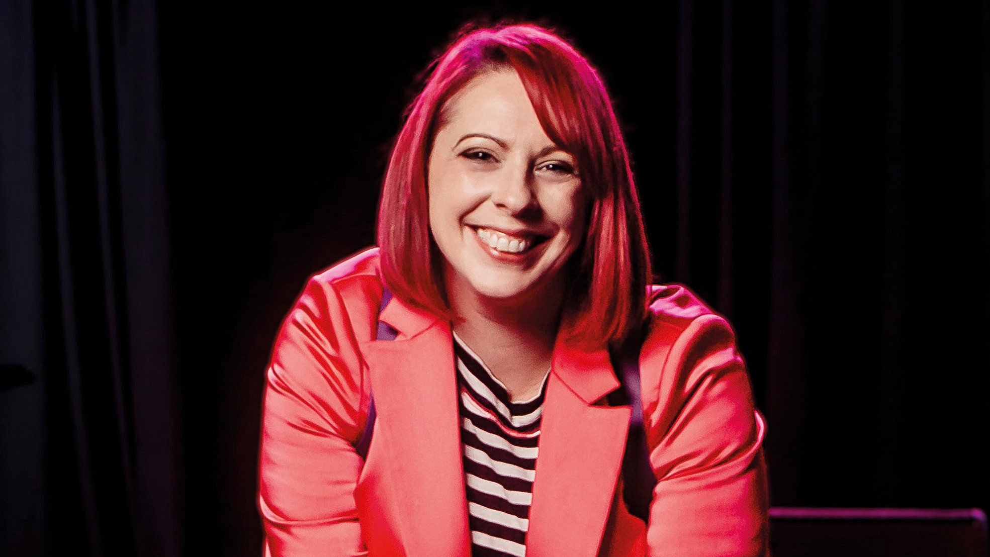 A woman (Carrie Davenport) in a bright pink blazer sits confidently with a camera, ready to capture moments in a dimly lit venue