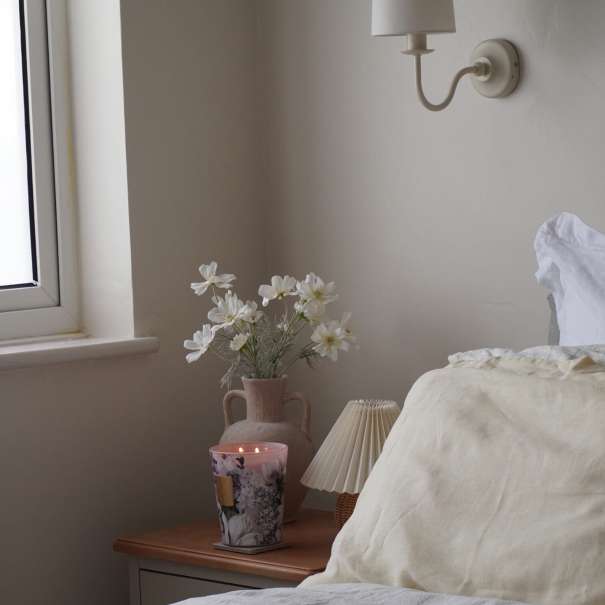 Neutral bedroom with lit floral candle, small table lamp and vase of white flowers on bedside table