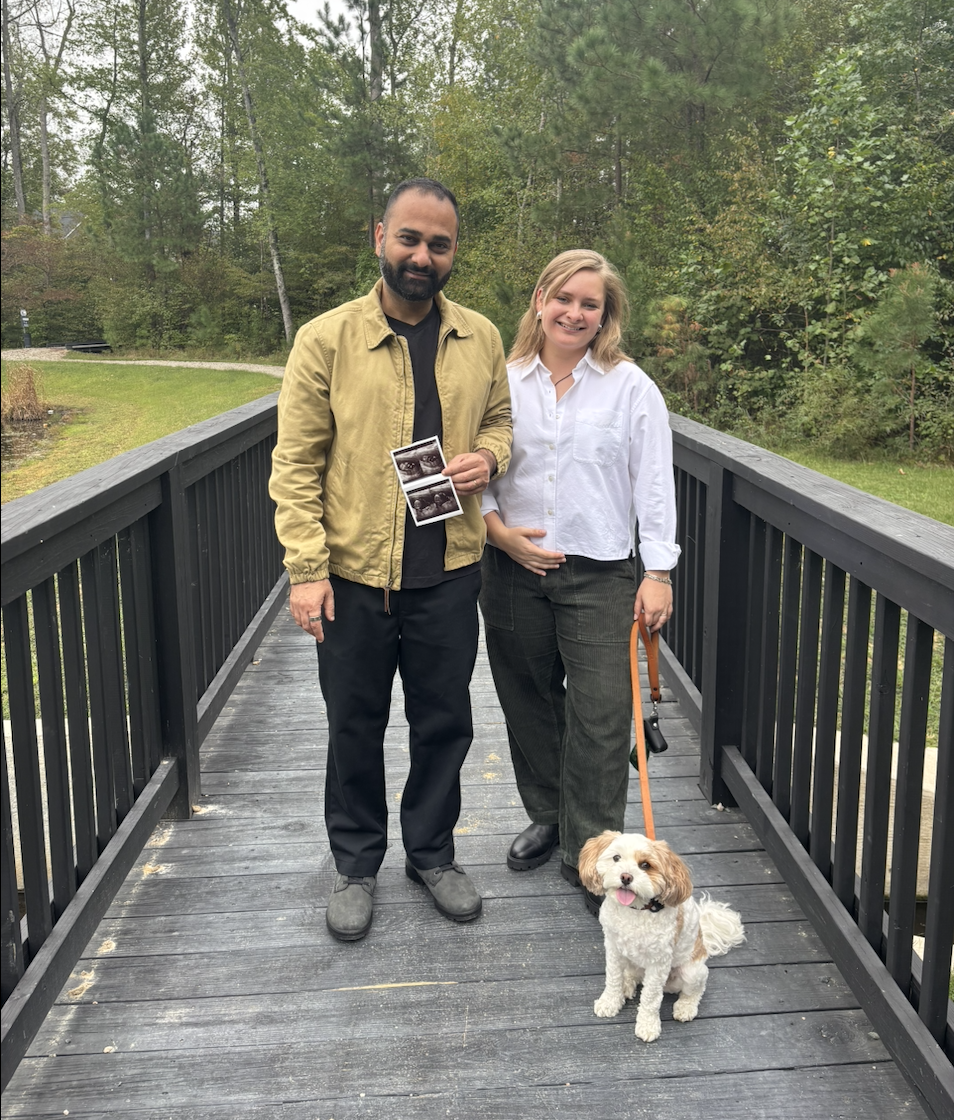 Lisa Vedernikova Khanna and her husband standing on a wooden bridge with their dog; her husband holds ultrasound images.