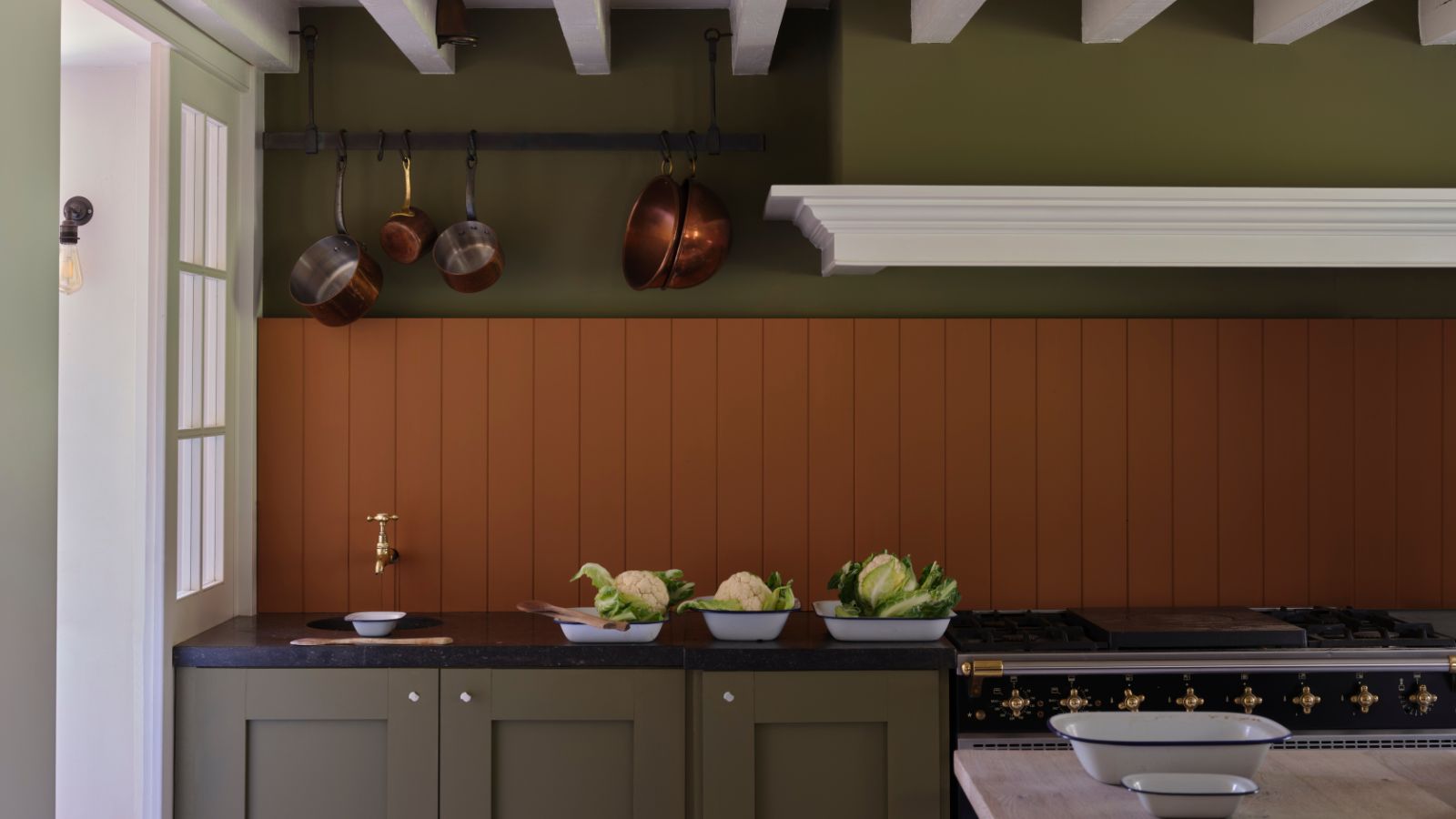 Dark green kitchen with dark orange wall panels and copper pans hanging on rail over a small sink. The counters are black, cabinets dark green, there is a window and door to the left, and shallow dishes with fresh produce by the sink
