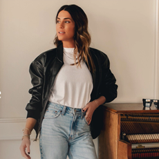 Woman wearing Abercrombie biker jacket, white tee, and blue jeans, leaning on piano