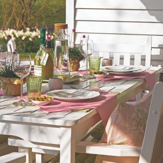 White wooden dining table and chairs in garden with white plates, pink placemats and glasses