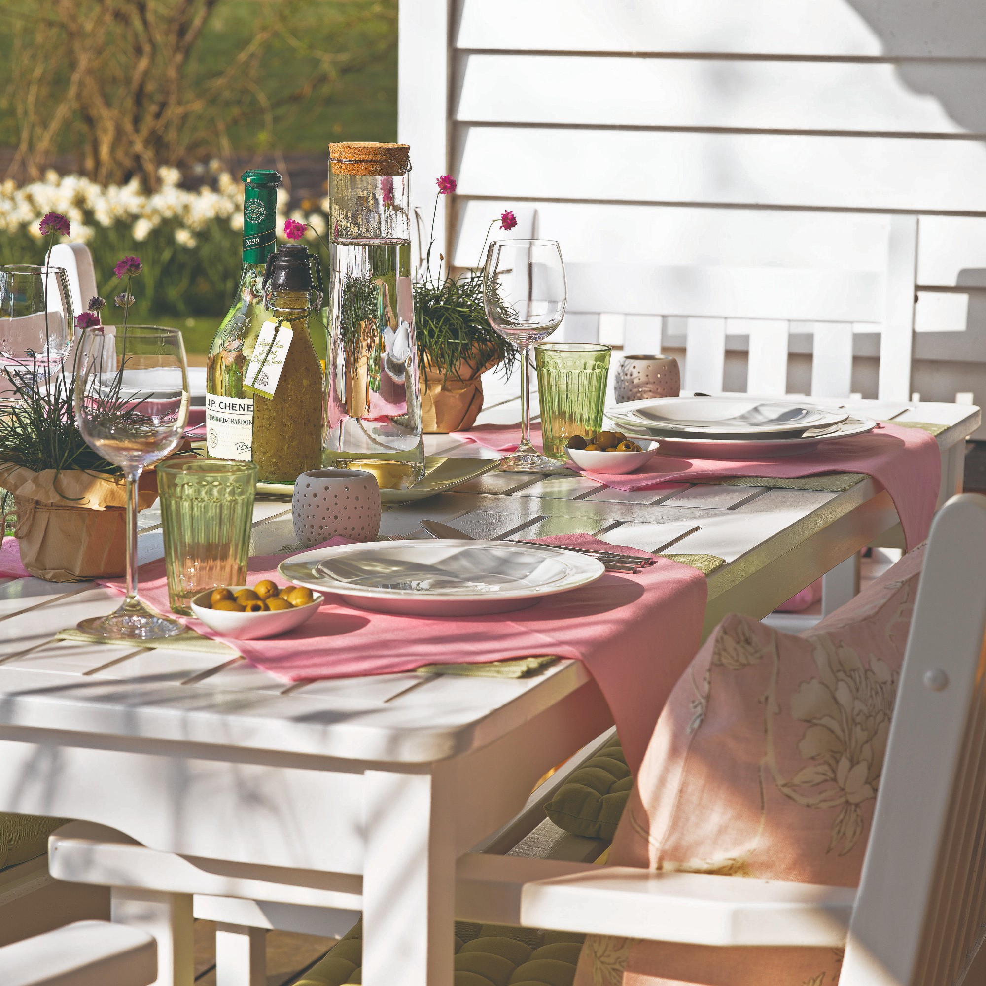 White wooden dining table and chairs in garden with white plates, pink placemats and glasses