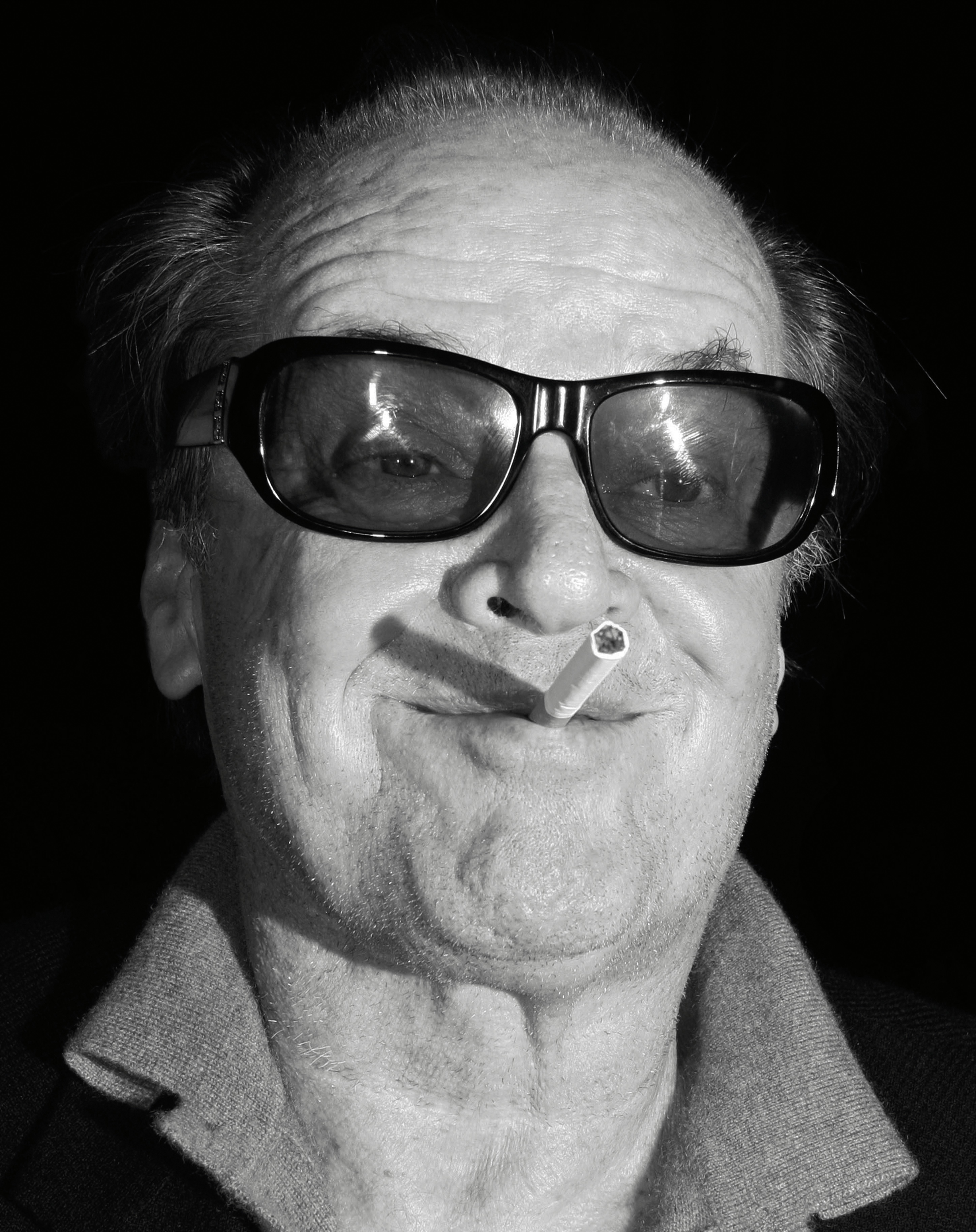 A close-up portrait of Jack Nicholson grinning with a cigarette in his mouth, wearing dark sunglasses.
