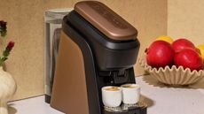 Close-up image of a brown Lavazza Tabli machine with two espresso shots on a white countertop with a beige bowl of pomegranates beside it.