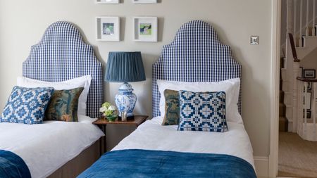 A cream bedroom with two twin beds with blue and white gingham headboards, white bedding, blue blankets, and matching patterned cushions. Between the beds is a wooden bedside table with a large white lamp with blue patterning and blue lampshade, with flowers beside it. Above the beds are small square, framed artworks, and to the right is an open doorway with a staircase seen beyond.