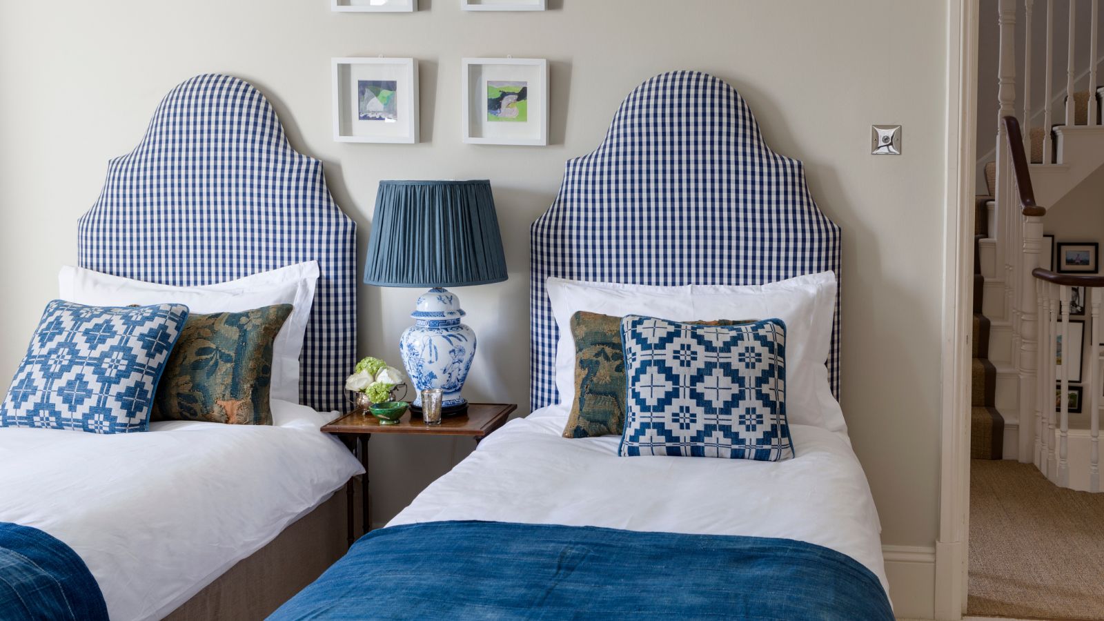 A cream bedroom with two twin beds with blue and white gingham headboards, white bedding, blue blankets, and matching patterned cushions. Between the beds is a wooden bedside table with a large white lamp with blue patterning and blue lampshade, with flowers beside it. Above the beds are small square, framed artworks, and to the right is an open doorway with a staircase seen beyond.