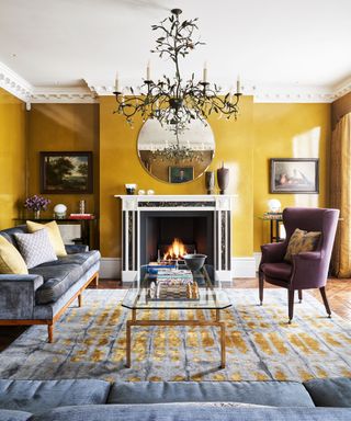 Yellow living room paint ideas with a white ceiling and cornicing, statement fireplace , large chandelier and blue-gray furniture.