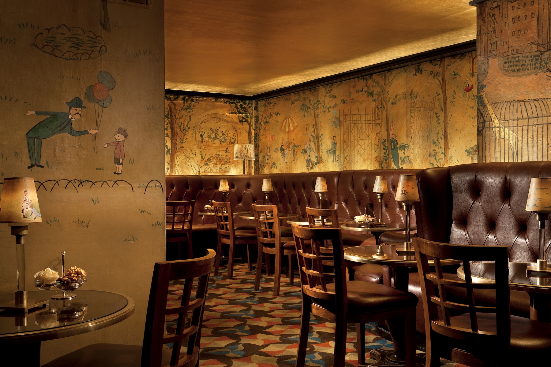 A golden-lit, spirited bar in New York features expansive handmade murals depicting everyday scenes in a cartoony style alongside leathery banquettes, wooden table sets, and similarly decorated table lamps.