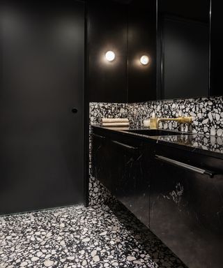 black bathroom ideas, black bathroom with terrazzo monochromatic floor and backsplash tiles with black cabinetry, black sink, brass faucet, black door