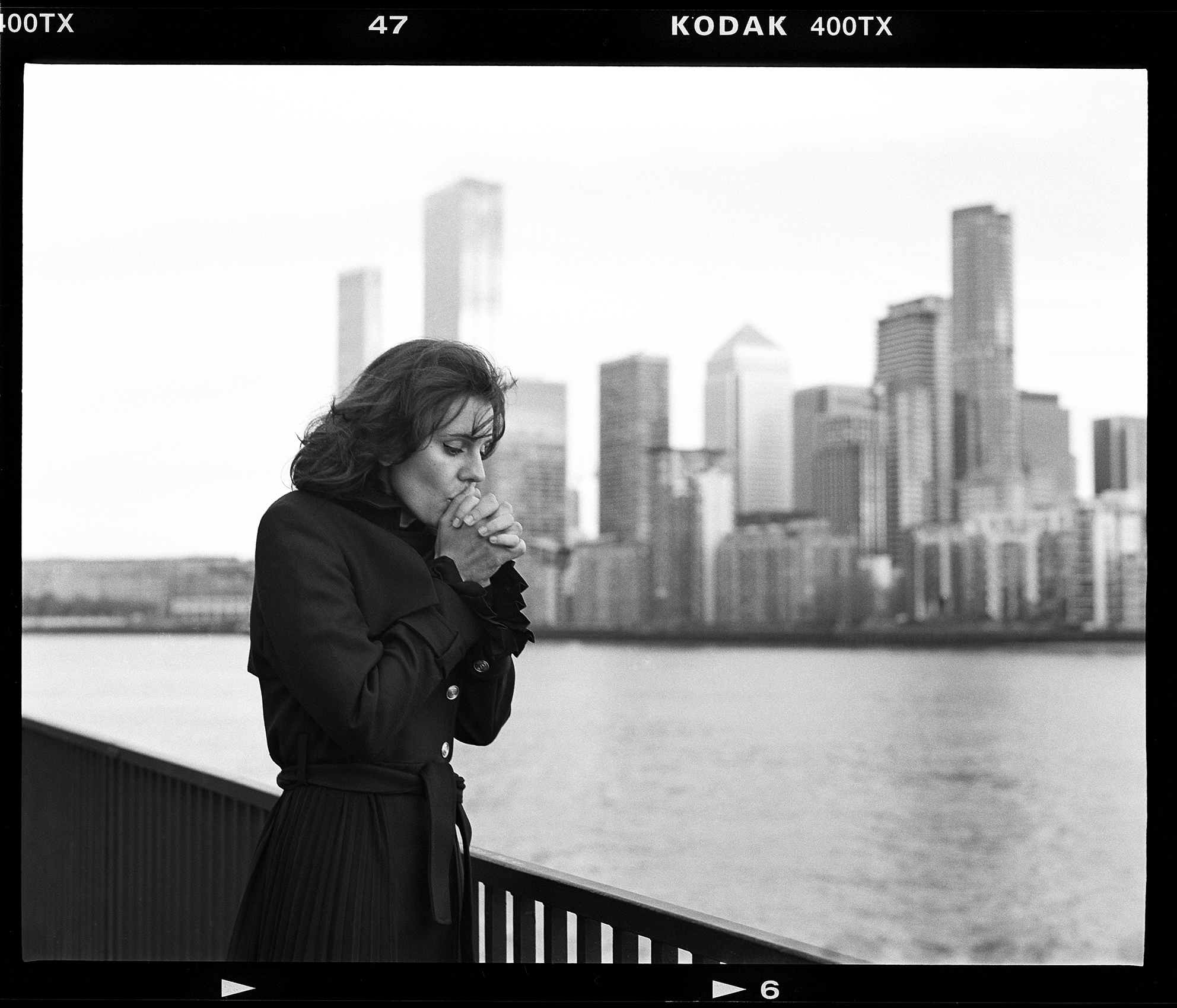 A figure in a black coat stands by a railing, overlooking a city skyline reflected in the water on a cloudy day