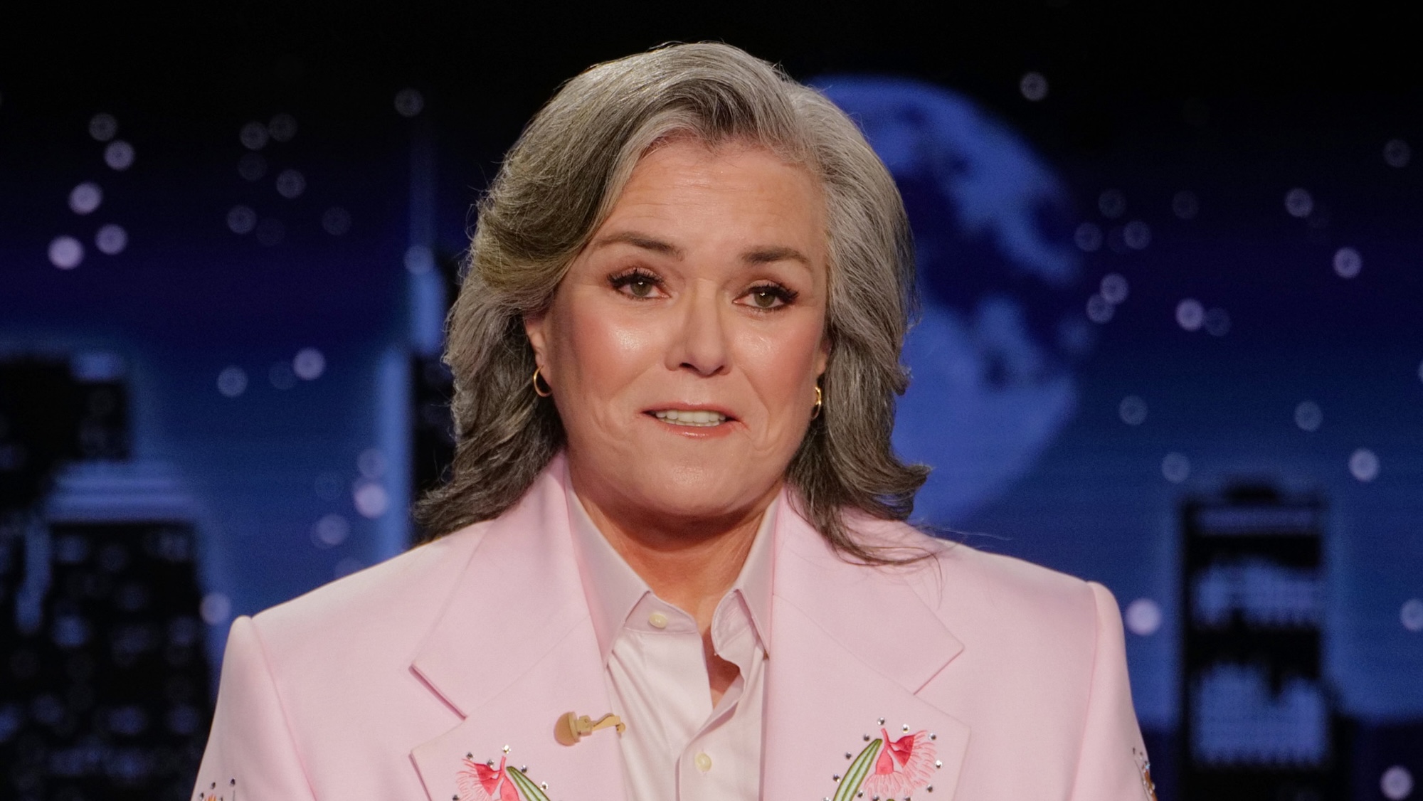 Rosie O'Donnell smirking at camera in pink jacket and white tie hosting Jimmy Kimmel Live! August 2026