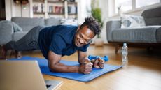 A man performs a plank exercise on a yoga mat in a living room. His forearms and toes are on the floor while he holds the rest of his body straight, elevated off the ground. His head is turned to look at a laptop screen next to him. Behind him we see a water bottle, light dumbbell weights, a pair of couches and a book case.