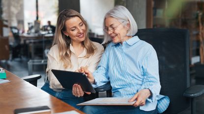 An older woman and her adult daughter smile as they discuss business issues in the older woman's office.