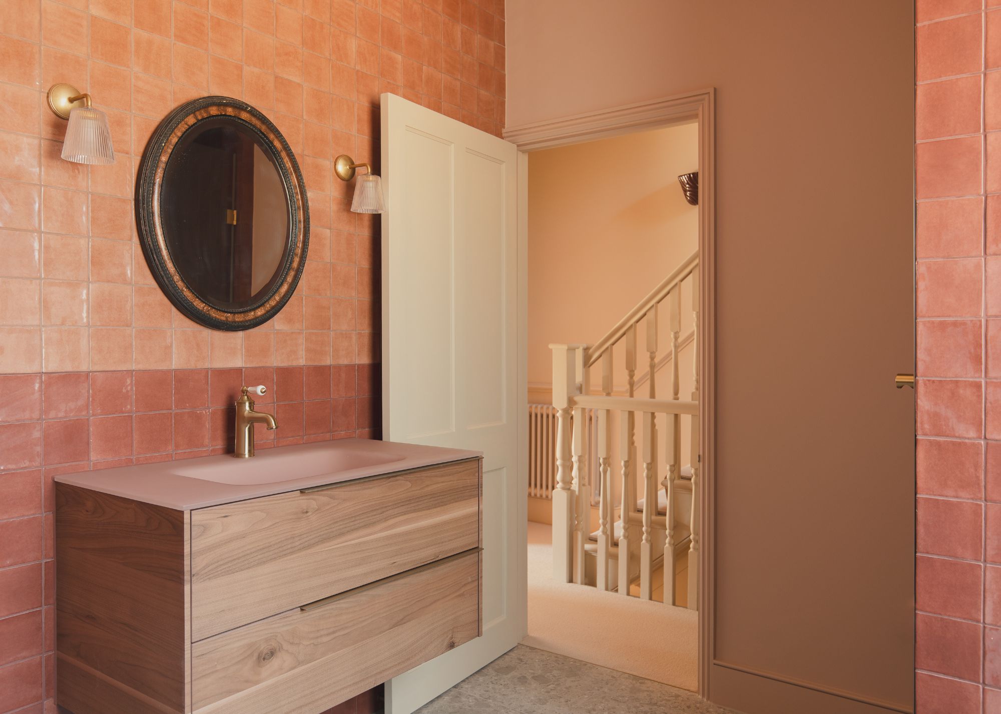 A modern bathroom decorated with earth tone colors with an accent wall of orange and clay tiles behind a floating vanity with a pink sink and round mirror above with a wall sconce either side