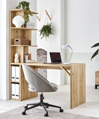 Wooden desk and shelf combo by Next, in an all-white office with a brick wall outside, and a gray desk chair on wheels