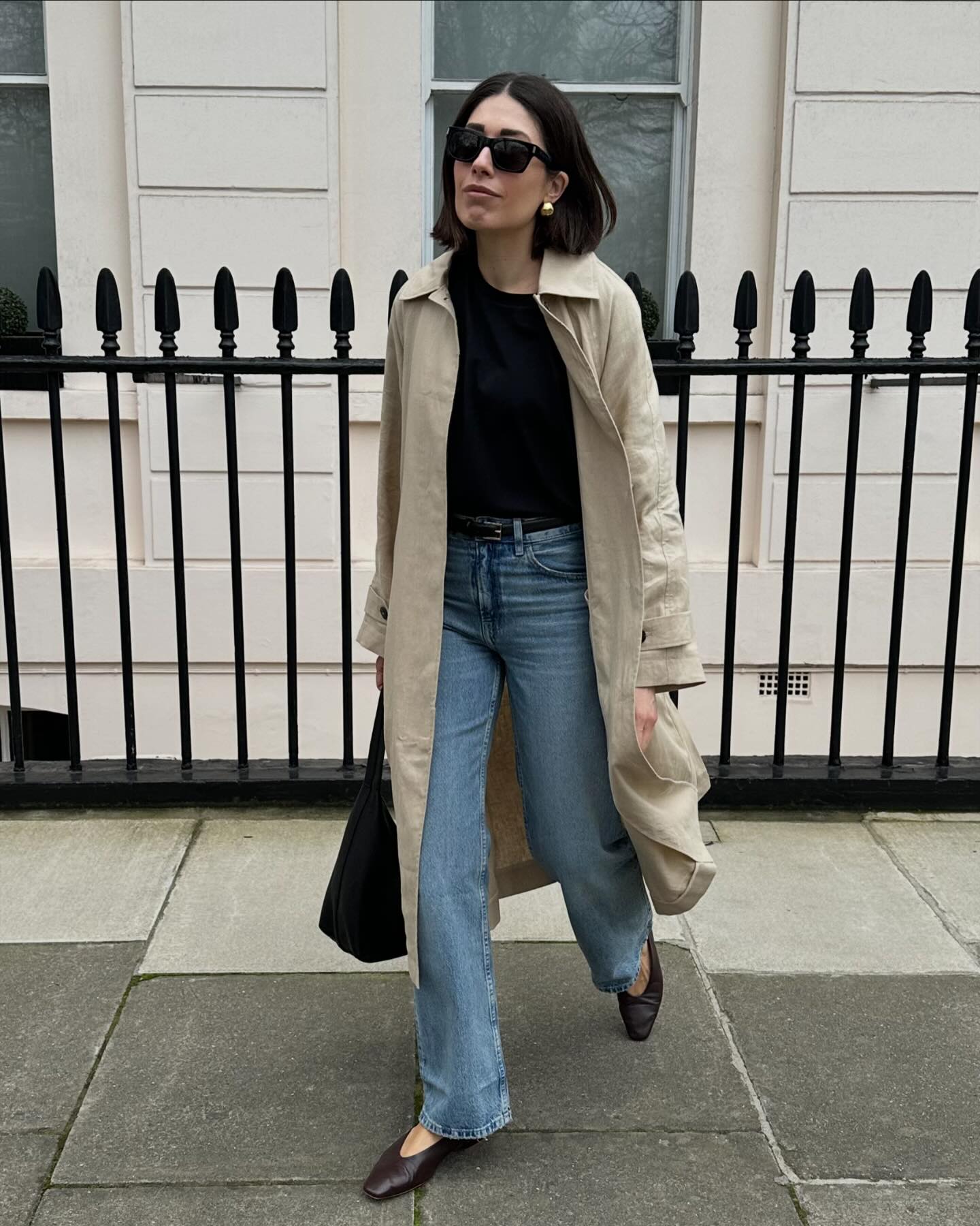 stylish influencer Lucy Alston walking on a sidewalk in London wearing a line trench coat, black top, straight-leg jeans, and brown flats