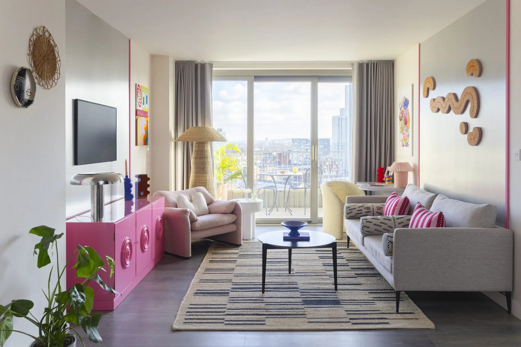 Quirky interior decorated with a pink lacquered console, rattan lamps and wall decorations, a softly shaped white armchair, and a beige sofa, along with plants and collectibles.