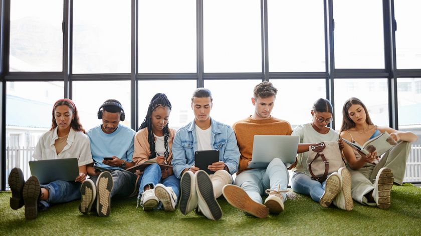 A group of young workers aka digital natives sat on the floor of an office against a large window, with devices such as laptops, tablets, and phones in their laps.