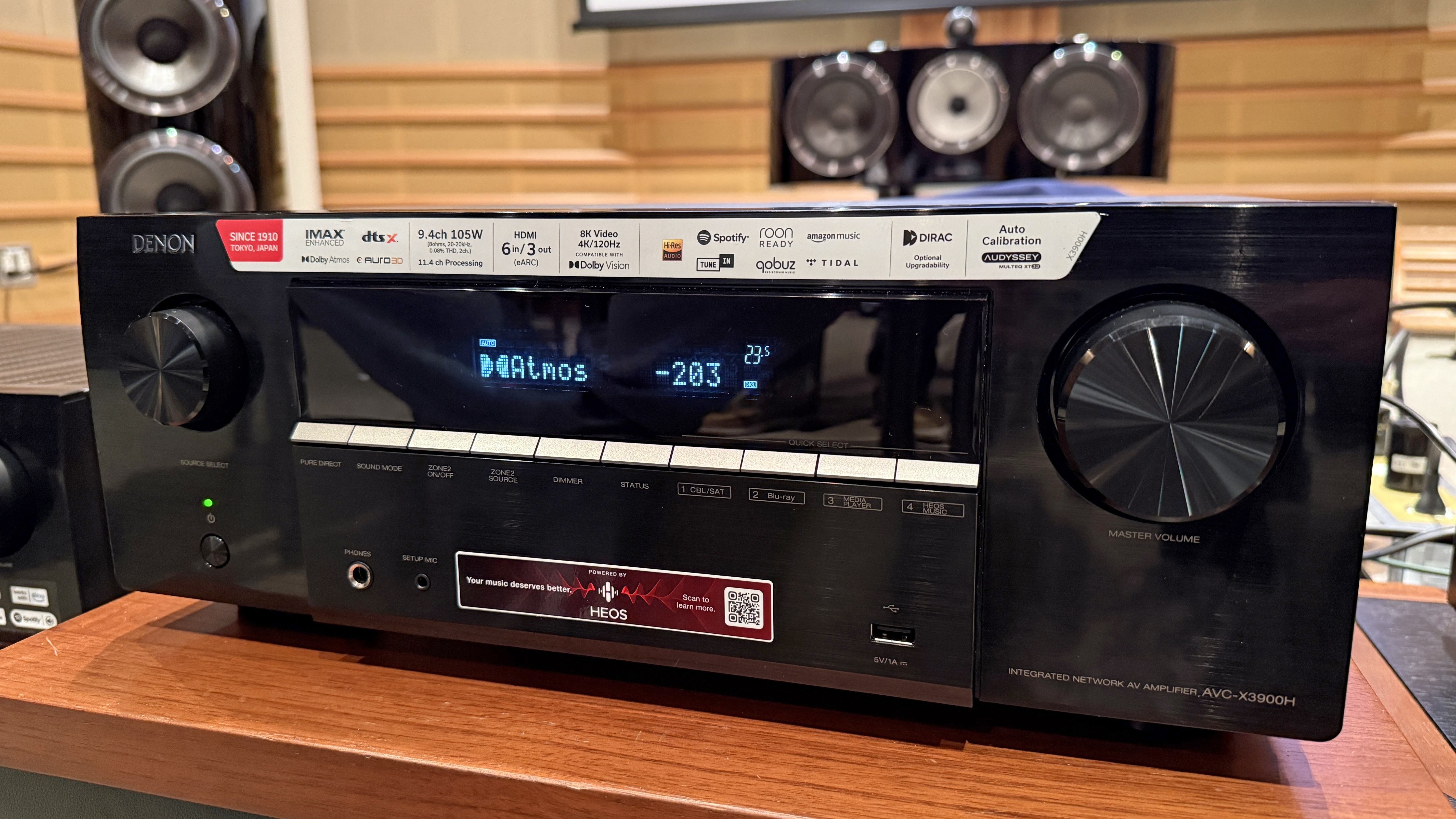 The Denon AVR-X3900H with Bowers &amp;amp; Wilkins speaker just visible behind it. It's a black box with some dials and buttons on the front. The screen says it's playing a Dolby Atmos feed