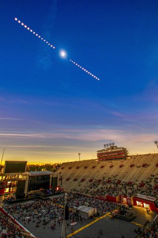 An eclipse time-lapse composite image shows the progression of a solar eclipse, framing totality in the middle in a darkening sky above a sparsely attended football stadium at Indiana University.