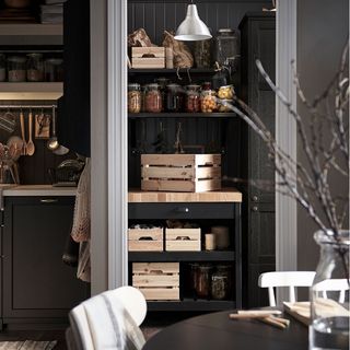 a black and grey kitchen with a black and wooden portable kitchen trolley inside a larder area, with wooden crates and jars of produce