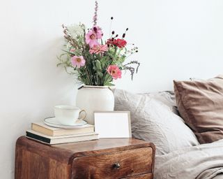 dark brown wooden nighstand with flowers and a pile of books and a white wall behind it and a bed next to it