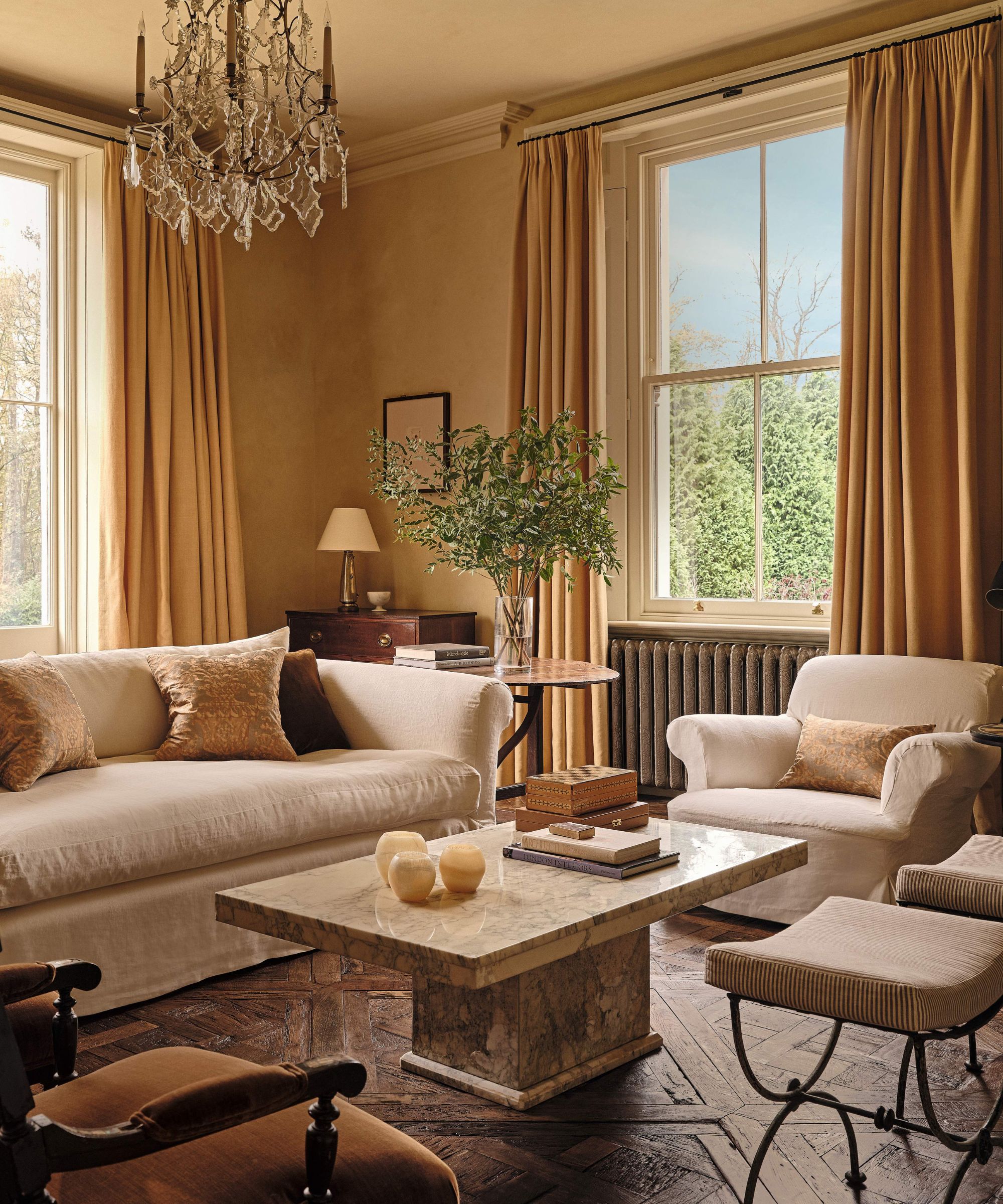 a warm deep yellow living room with grand georgian windows, neutral slip covered sofa and armchairs, marble coffee table, antique stools and chandelier