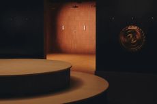 A dimly lit sauna room with a curvilinear, central podium in muted brown, a brass, low-relief wall decoration, and red brick-wall showers.
