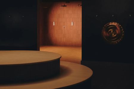 A dimly lit sauna room with a curvilinear, central podium in muted brown, a brass, low-relief wall decoration, and red brick-wall showers.