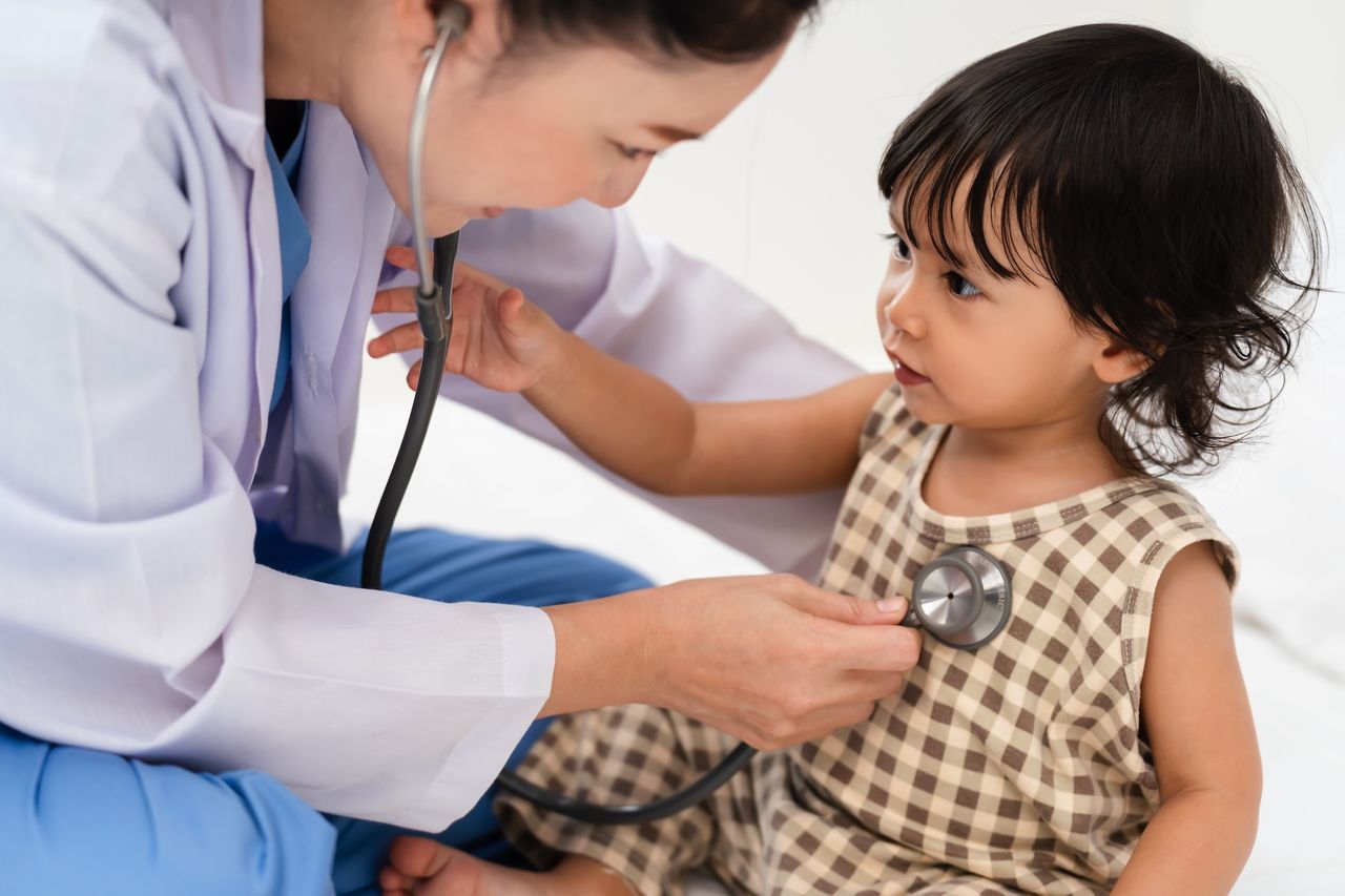 A woman wearing a lab coat holds a stethoscope to a toddler's chest.