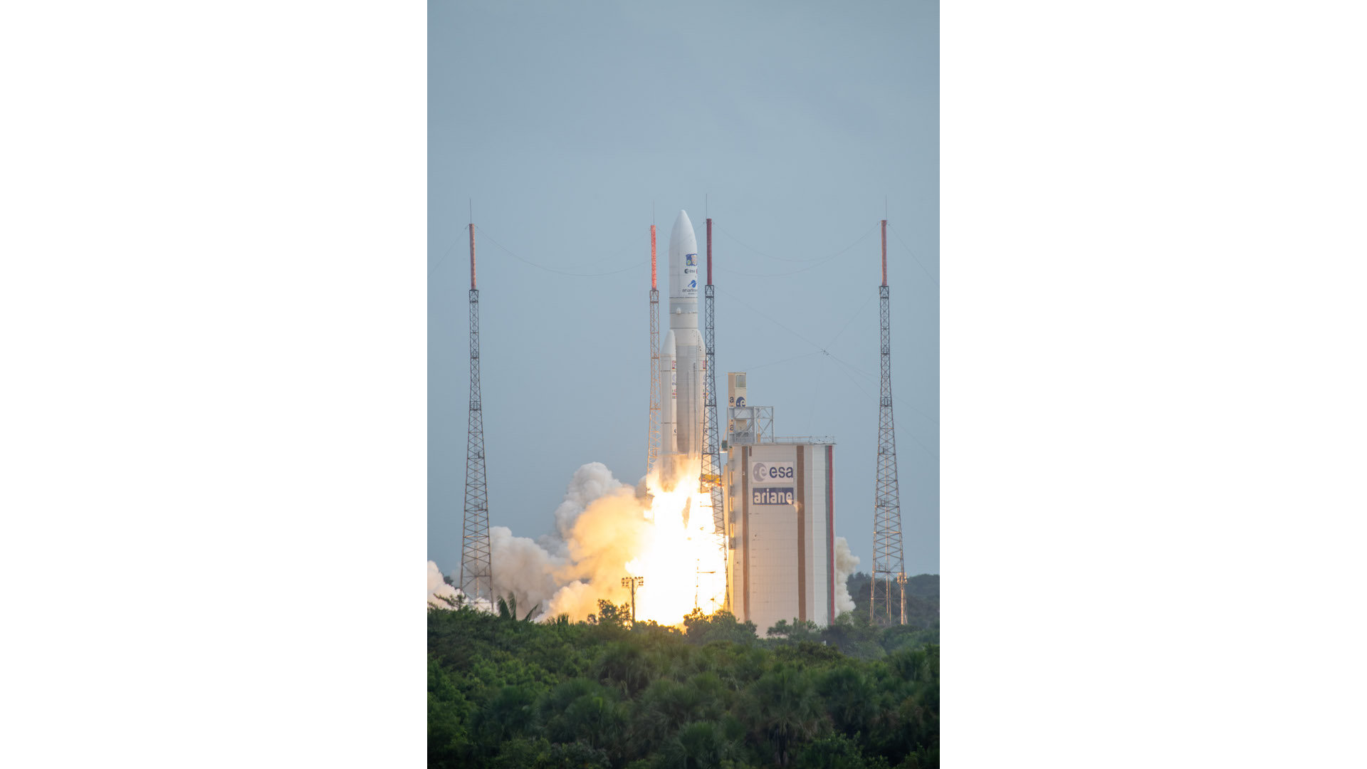 An Ariane 5 rocket lifts off against a grey/blue sky. The yellow/orange from the engine flames reflects against the launch tower. A green expanse of forest sits in the foreground.