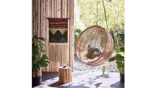HK Living Natural Rattan Hanging Egg Chair in a sunny outdoor space with a rattan wooden wall and a plant next to it