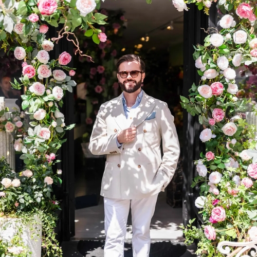 Image of a man in a white suit and sunglasses standing under an archway of white and pink roses.