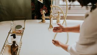 female sat on edge of rolltop bath with hands under water running from gold tap