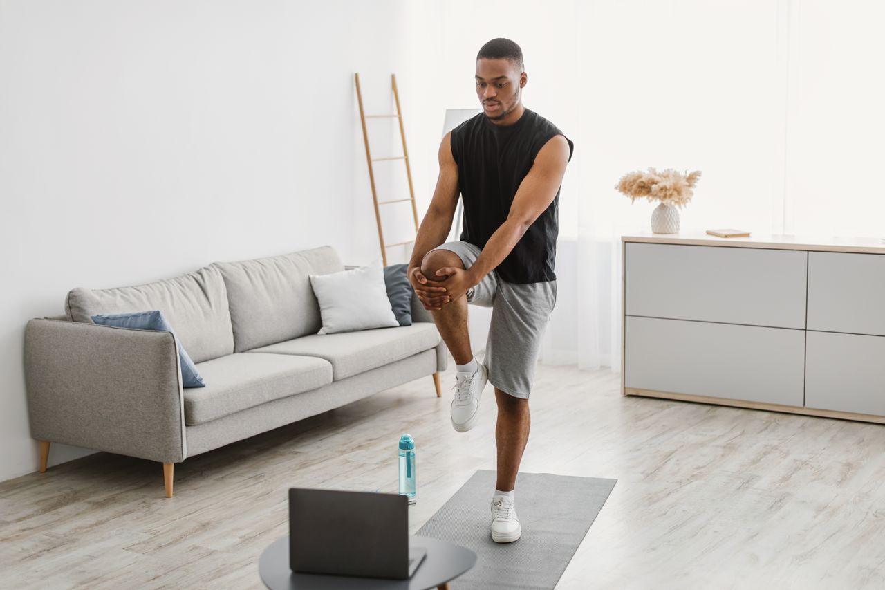 A man in shorts, vest and sneakers stretches his leg while standing on a yoga mat in a living room looking down at an open laptop. He is balanced on this left leg, while his right leg is elevated and bent at the knee; both hands clasp the knee. Behind him is a couch, decorative ladder, chest of drawers and a vase full of flowers.